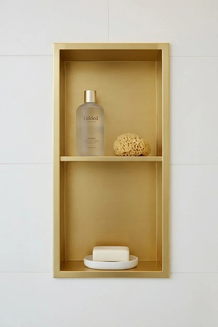 A gold-framed two-tier wall-mounted shelf with a matte gold interior, styled with a frosted bottle, a sponge, and a bar of soap on a white tiled wall.