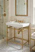A white ceramic bathroom sink with a polished gold metal pedestal stand, set in a classic bathroom with marble tile, a gold-framed mirror, and a glass shower enclosure.