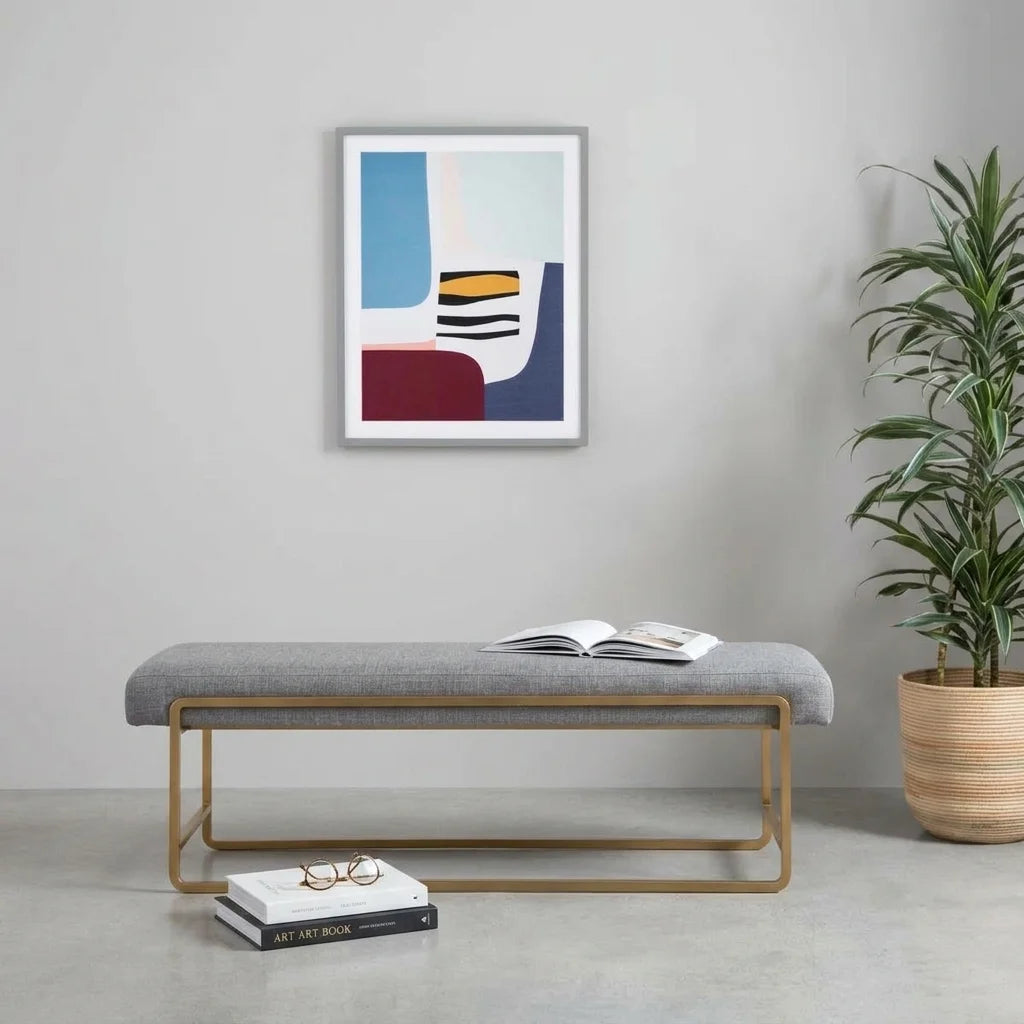 A modern gray upholstered bench with a gold metal frame, styled in a minimalist room with an abstract art print on the wall, an open book on the seat, glasses on a book, and a potted plant to the side.