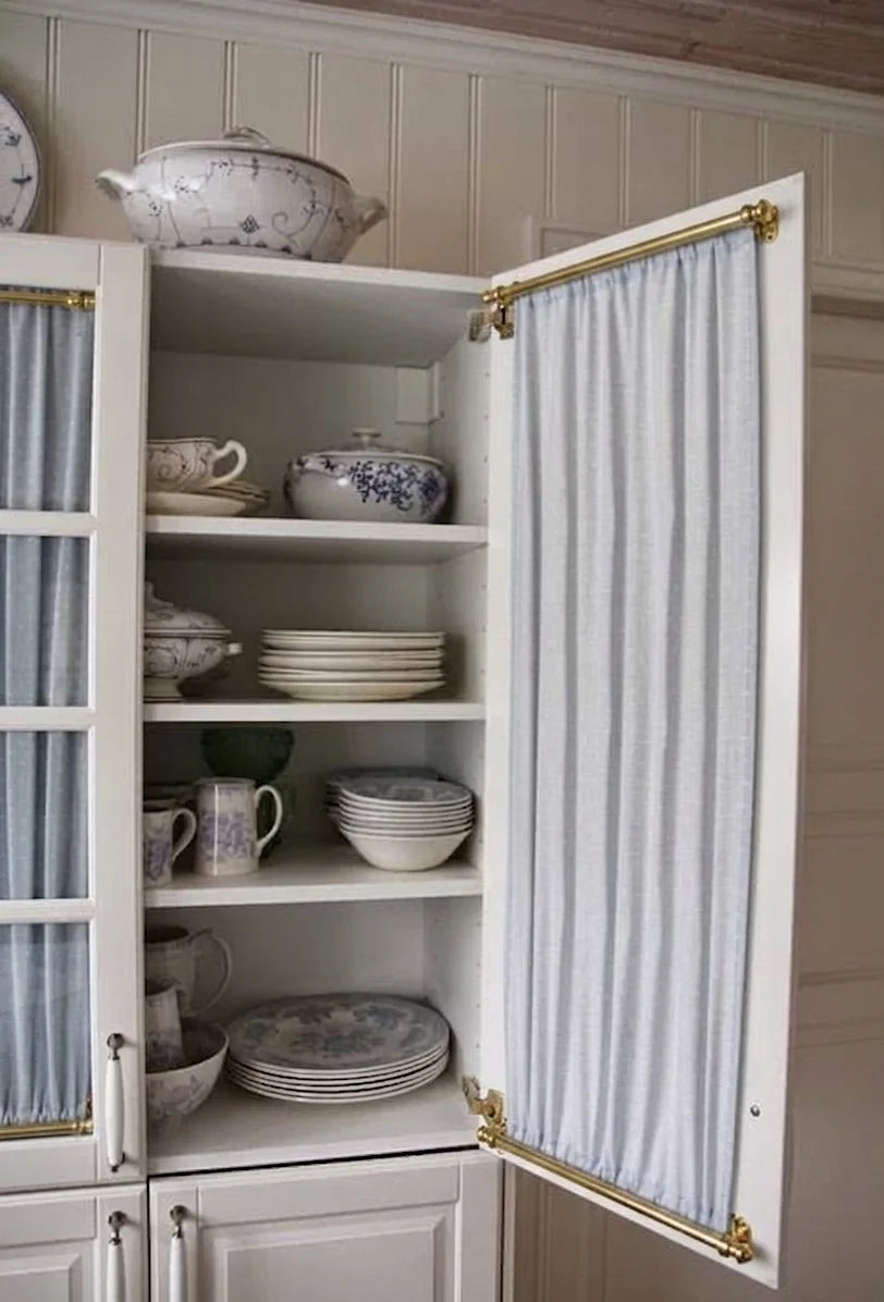 A white kitchen cabinet with brass hardware, shown open to reveal shelves of dishes inside. The cabinet door is covered with a light blue and white striped fabric panel, and a matching striped curtain hangs on the left side.