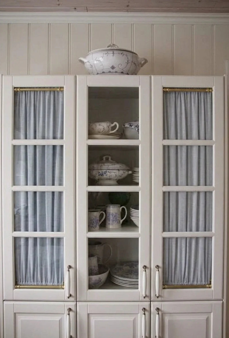 A tall white china cabinet with glass doors and light blue pleated curtains, displaying ceramic dishes and a teapot inside, set against a beige paneled wall.