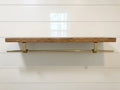 A floating wooden shelf with a natural oak finish and a brass metal rod mounted beneath it, shown against a white shiplap wall.