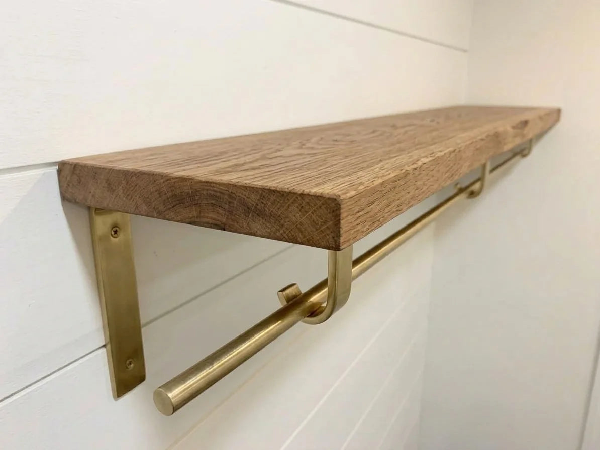 A rustic wooden floating shelf with a natural oak finish, mounted on a white shiplap wall with a brushed brass metal rod and bracket underneath.