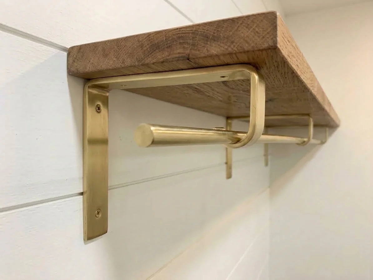 A rustic wooden floating shelf mounted on a white shiplap wall, supported by a brushed brass metal bracket with an integrated towel bar.