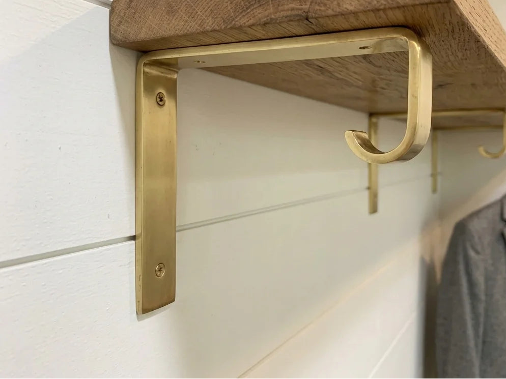 A rustic wooden shelf mounted on a white shiplap wall with brass metal brackets and hooks, shown with a gray blazer hanging on a hanger in the background.