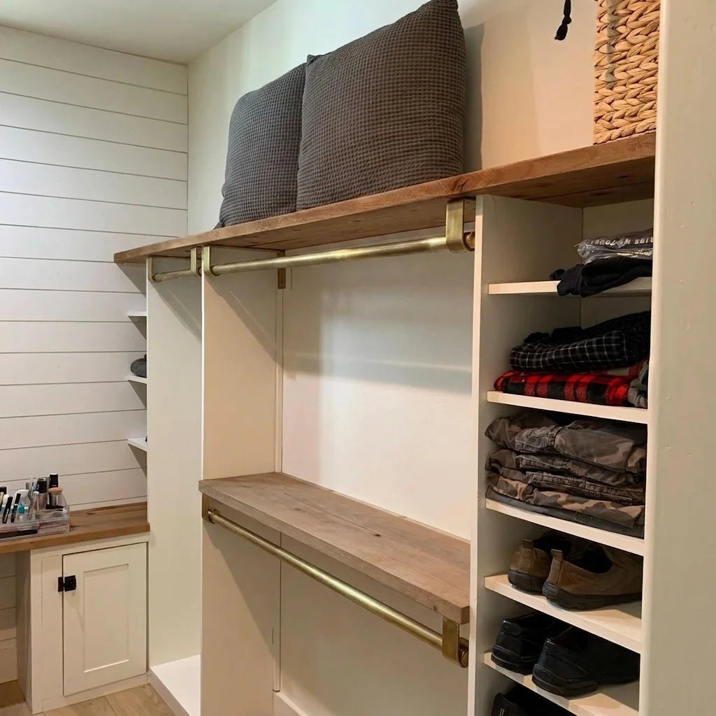 A built-in closet organizer with open shelving, two wooden shelves, brass hanging rods, folded clothing on the side shelves, two gray pillows on top, and a small vanity cabinet on the left.