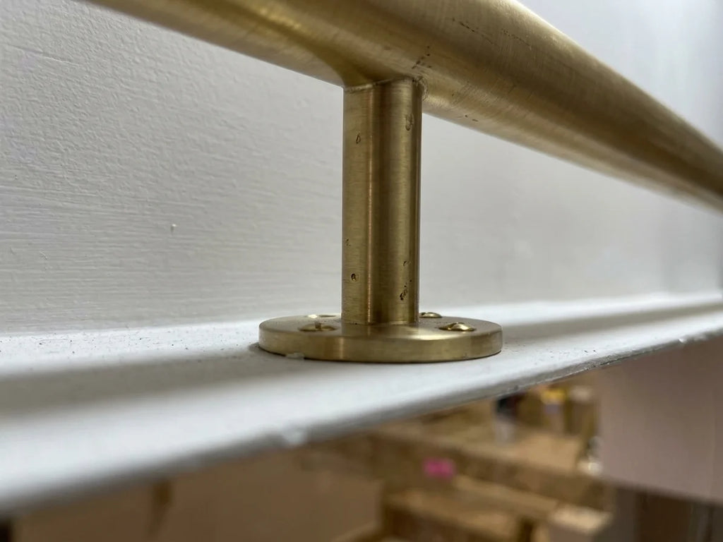 A close-up of a brushed brass or bronze metal handrail mounted to a white wall and white ledge, with a circular base plate visible.