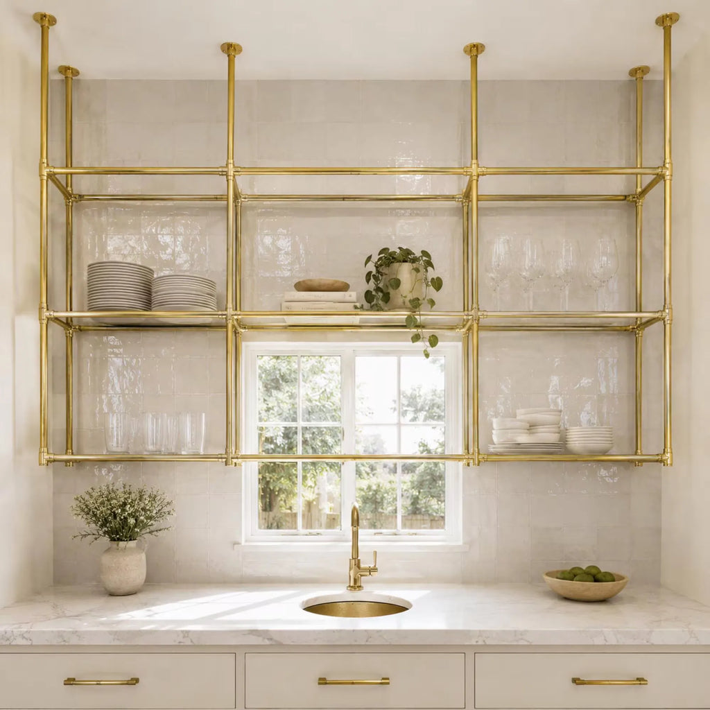 A modern kitchen scene with a large brass wall-mounted shelving unit above a white marble countertop and sink. The open shelves are styled with white plates, glassware, books, and a small potted plant.