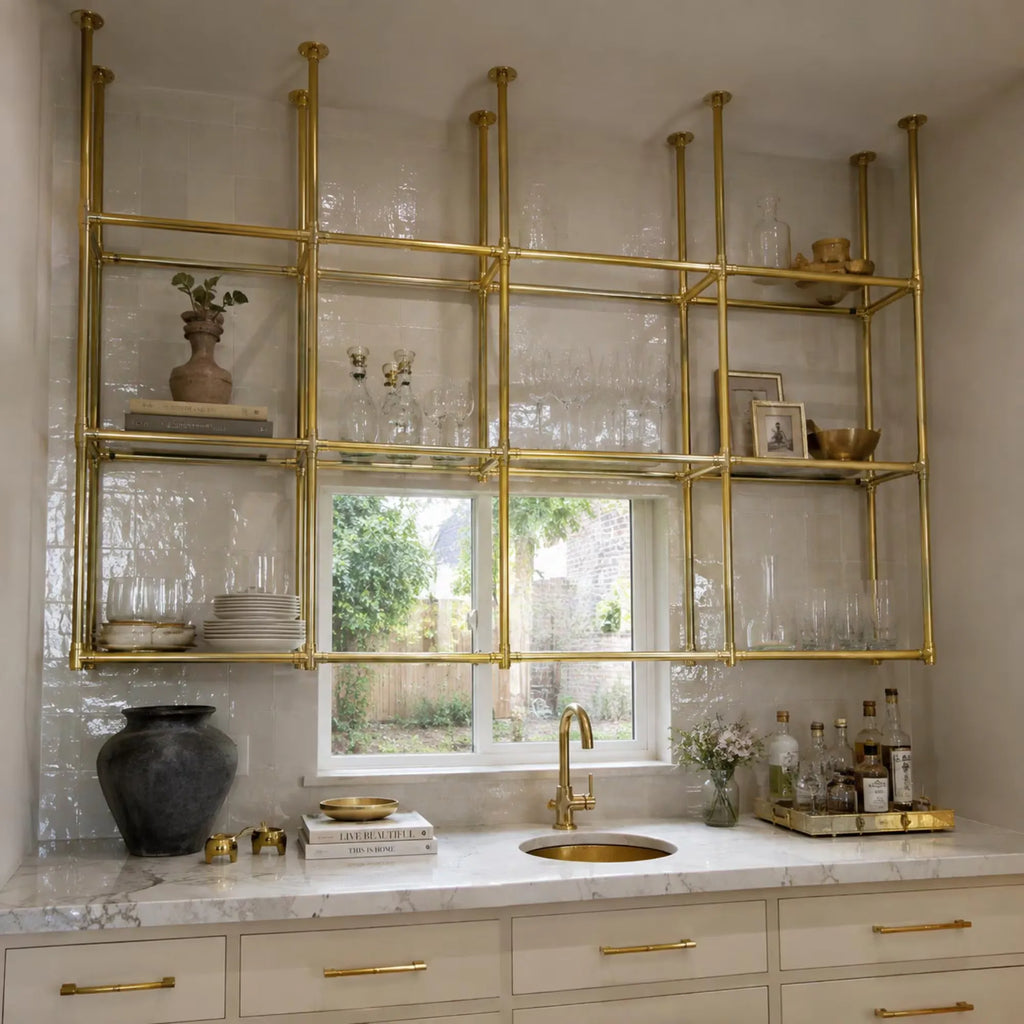 A modern kitchen featuring a large brass open shelving unit mounted on a white tiled wall above a marble countertop with a gold faucet and sink.