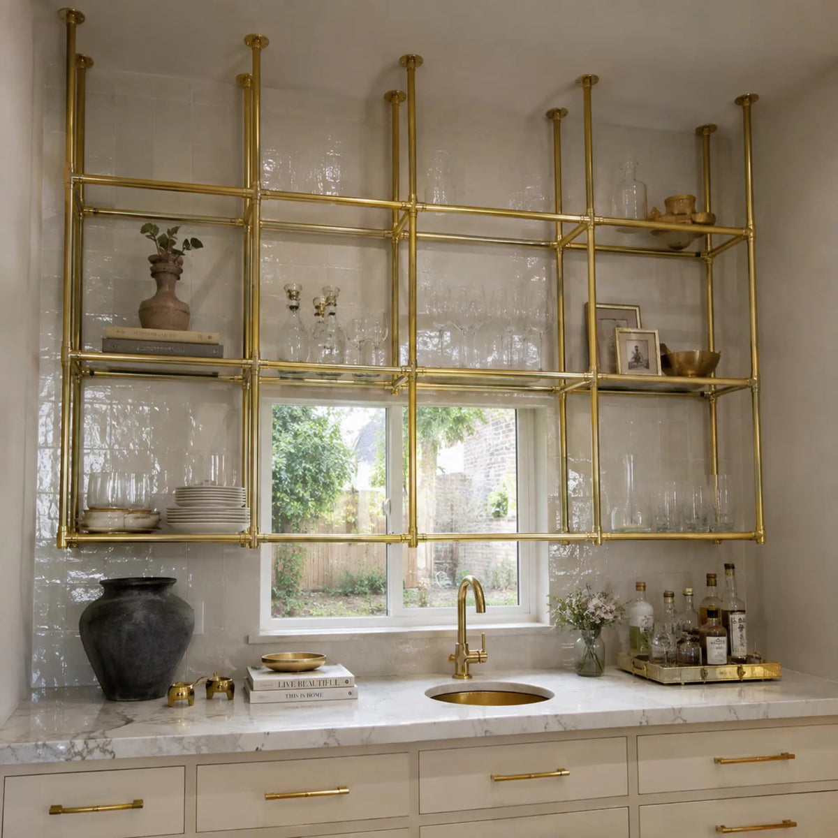 A modern kitchen featuring a large brass open shelving unit mounted on a white tiled wall above a marble countertop with a gold faucet and sink.