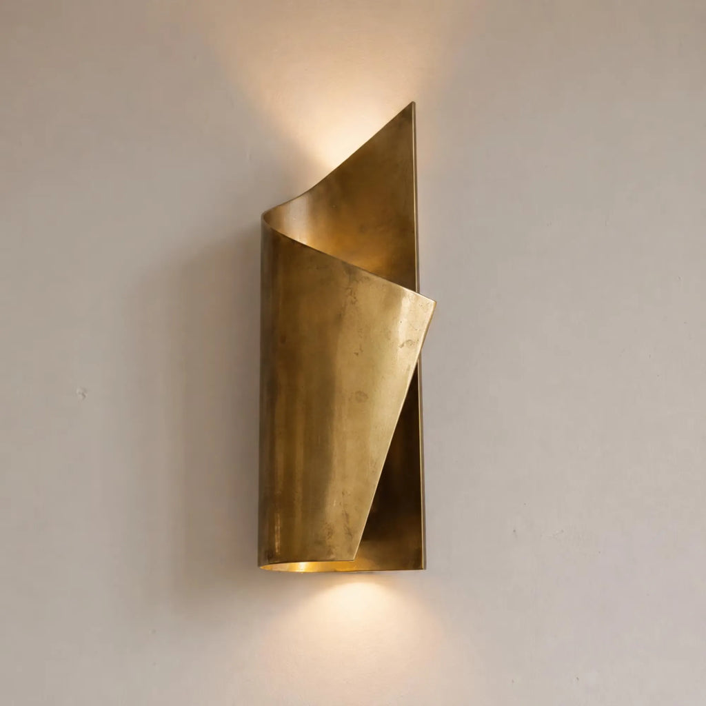 A modern wall-mounted sconce with a sculptural, twisted triangular design in brushed brass, casting a warm glow against a neutral off-white wall.