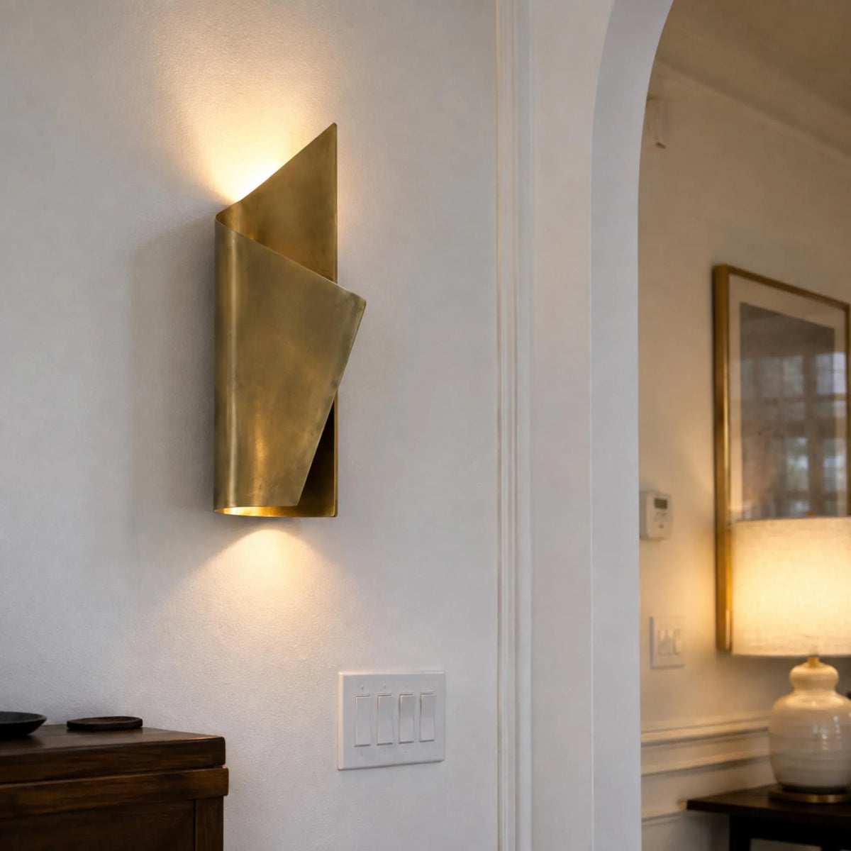 A modern wall-mounted sconce with a folded, geometric bronze metal design, illuminated on a textured off-white wall in a home interior setting.