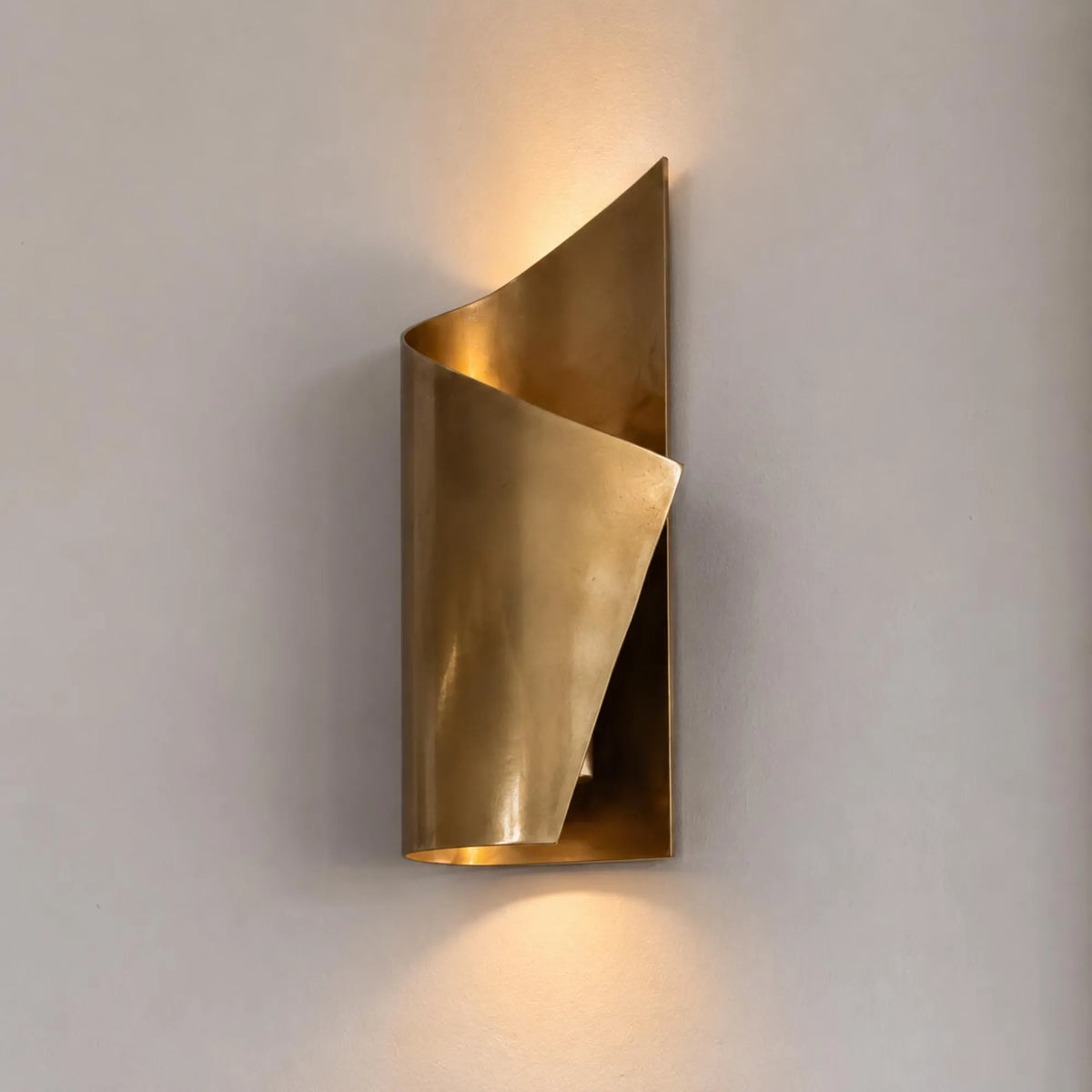 A modern wall-mounted sconce with a sculptural, curled metallic gold design, casting a warm glow against a neutral light gray wall.