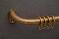 A close-up of a curved brass shower or towel bar mounted on a dark textured wall, with four hanging rings attached.