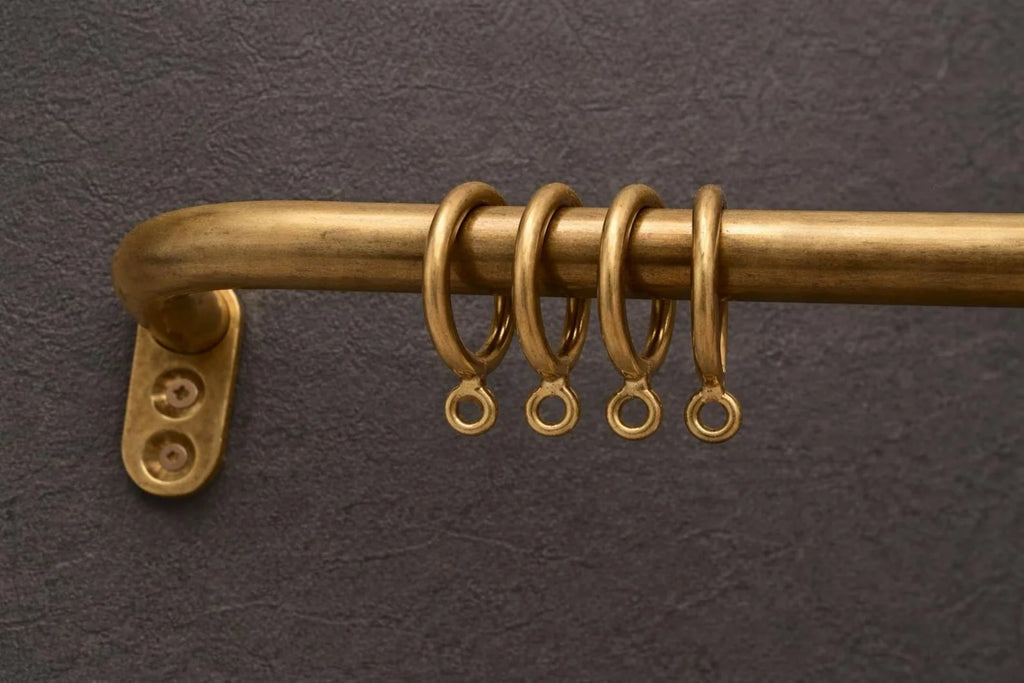 A close-up of a brass curtain rod with a wall mount bracket and four matching brass curtain rings hanging from it, set against a dark textured background.