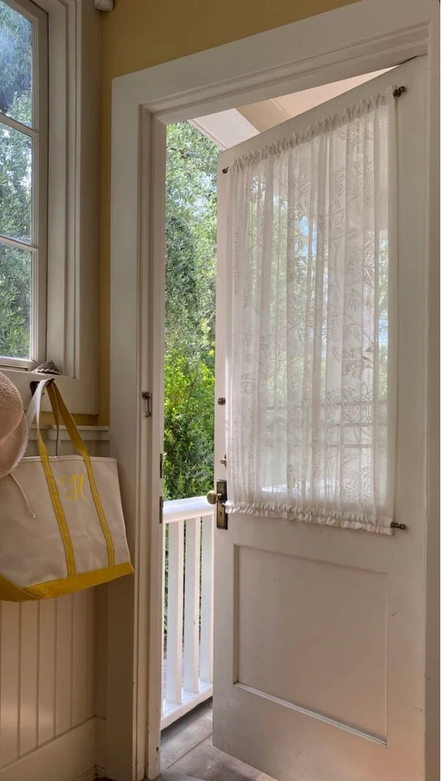 A sheer white embroidered curtain panel hanging on a white front door in a bright entryway, with a window, wall hooks, a hat, and tote bag visible on the left.