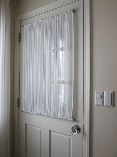A white sheer curtain panel hanging on a silver metal rod over a white paneled door, shown in a home interior setting with beige walls and a light switch visible.